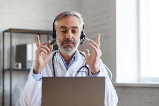 Ritratto di medico maschio anziano dai capelli grigi nel suo ufficio utilizzando laptop per chat video con un paziente. consultazione online con il medico per diagnosi e raccomandazione di trattamento. concetto di telemedicina.