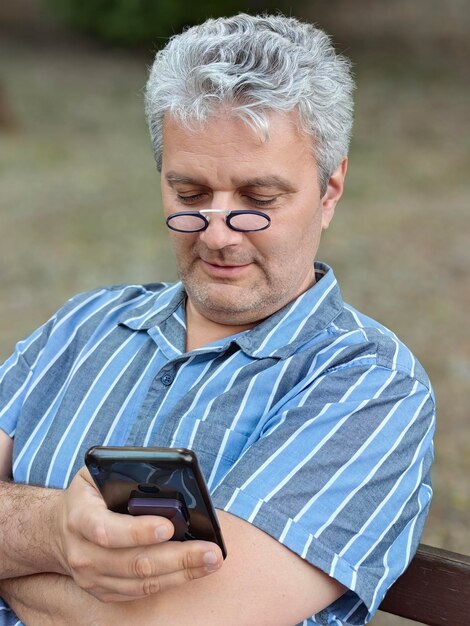 Photo portrait of an senior man sitting on a bench in a public park and using his smartphone