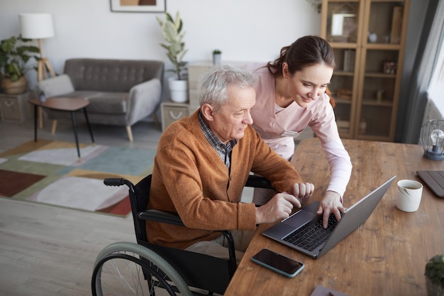 Portrait of senior man in wheelchair using laptop at retirement home with nurse assisting him, copy space