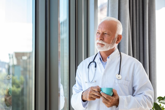 Портрет старшего зрелого профессионального врача со стетоскопом, держащего синюю чашку кофе