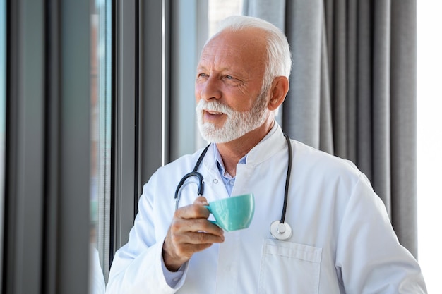Portrait of senior mature health care professional doctor with stethoscope holding blue cup of coffee