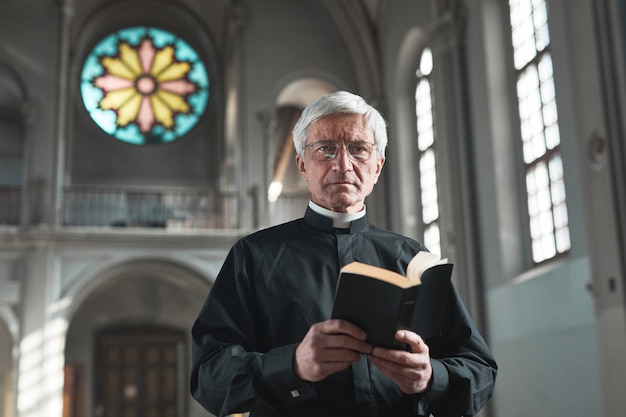 Foto ritratto di un sacerdote anziano che tiene in mano la bibbia e guarda la telecamera mentre si trova in chiesa