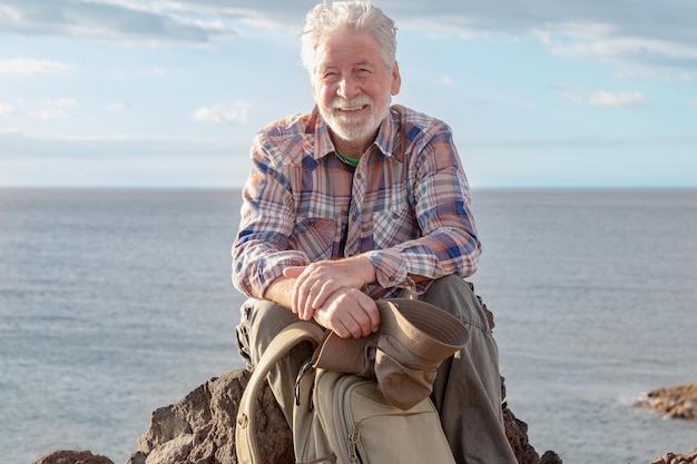 Portrait of senior smiling man resting in outdoor excursion at sea sitting on the cliff looking at camera Mature attractive bearded man on a hiking trip enjoying freedom and healthy vacation