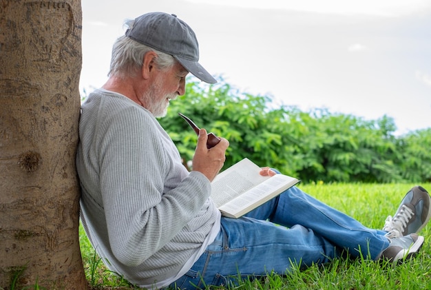 Portrait of senior smiling man sitting on the grass in public park having relaxed moments reading a book Caucasian elderly bearded male smoking pipe