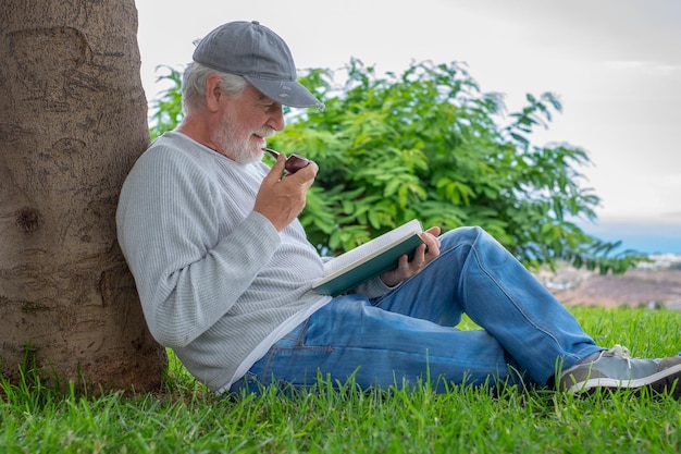 Portrait of senior smiling man sitting on the grass in public park having relaxed moments reading a book Caucasian elderly bearded male smoking pipe