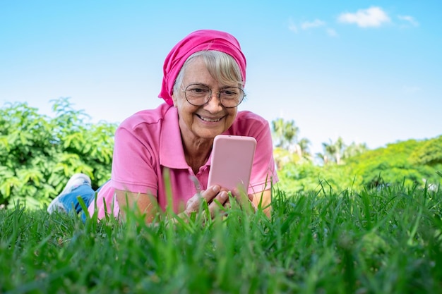 Portrait of senior smiling woman lying on the grass in public park using mobile phone with pink cover Romantic elderly lady enjoying free time and retirement