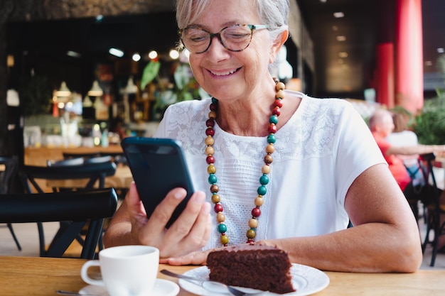 チョコレートケーキとコーヒーカップでコーヒーショップで休憩のためにリラックスしている年配の笑顔の女性の肖像画。携帯電話を使用して眼鏡とネックレスを持つのんきな年配の女性