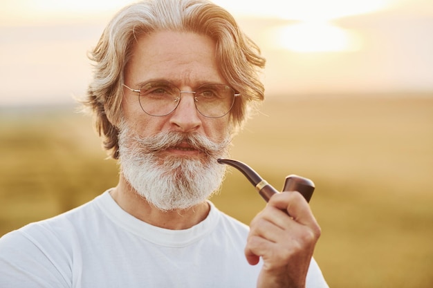 Portrait of senior stylish man with grey hair and beard that standing outdoors on field at sunny day and smoking