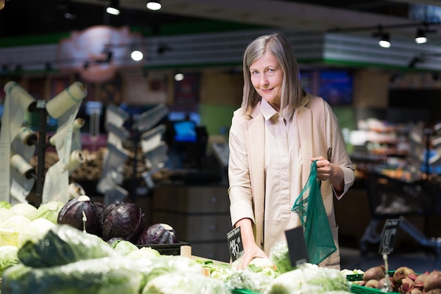 食料品店のスーパーマーケットの年配の女性の肖像画は、野菜を掘り、笑顔でカメラを見ながらエコバッグに入れます