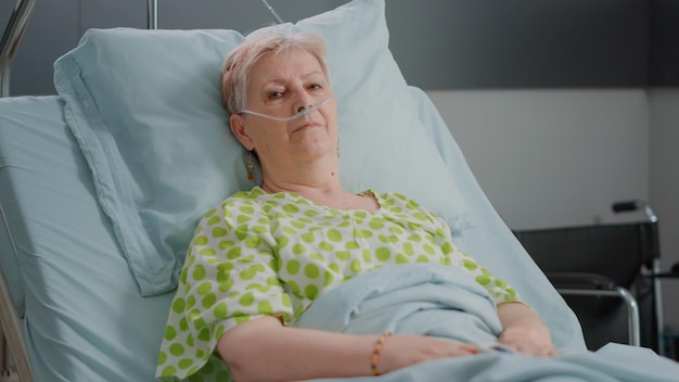 Portrait of senior woman laying in hospital ward bed with nasal\
oxygen tube and iv drip bag. hospitalized patient with sickness\
waiting to receive healthcare treatment and medical\
assistance.