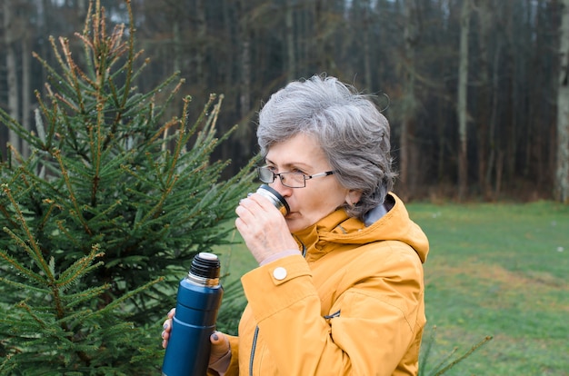 Portrait senior woman outside in the forest. Elderly woman drinking hot tea while walking. Concept hike, warming drink in cold weather.