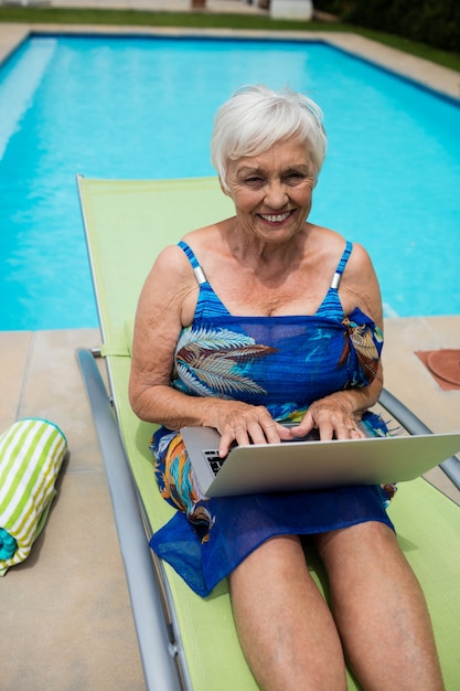 Ritratto di donna senior utilizzando laptop sulla sedia a sdraio a bordo piscina