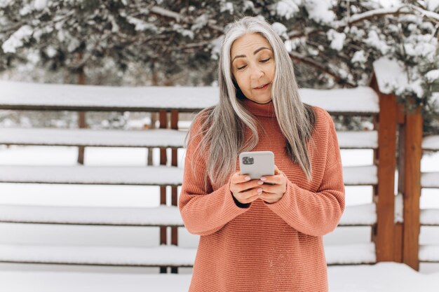 Портрет пожилой женщины с седыми волосами в вязаном свитере, разговаривающей по телефону на фоне снежной погоды зимой