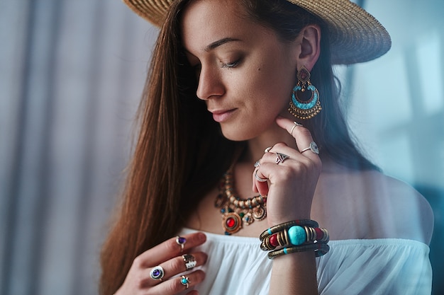 Ritratto di donna sensuale boho chic bruna che indossa camicia bianca e cappello di paglia con orecchini, bracciali, collana e anelli. vestito bohemien zingaro alla moda hippie con dettagli di gioielli