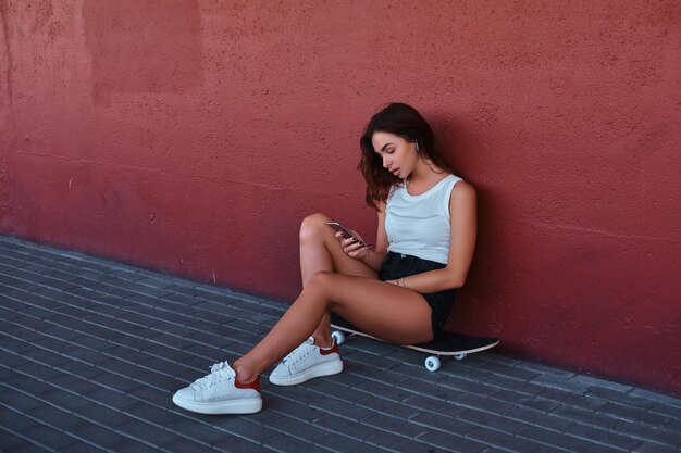Портрет чувственного хипстера, сидящего на скейтборде со смартфоном и слушающего музыку, опираясь на стену.