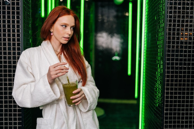 Портрет чувственной сексуальной молодой женщины в белом халате, держащей в руках стакан со свежевыжатым витаминным соком, стоящей и позирующей, глядя в сторону