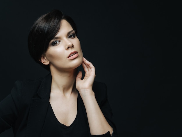 Portrait of sensual woman with short haircut and light makeup standing over black wall. Charming confident lady wearing black elegant clothes.