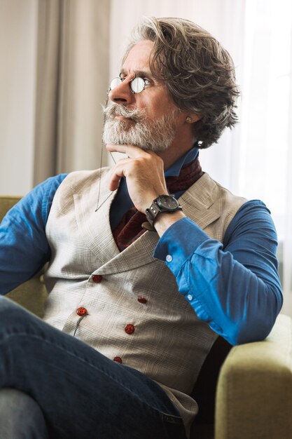 Portrait of serious adult professor man wearing stylish suit and eyeglasses looking aside while sitting on armchair in hotel apartment