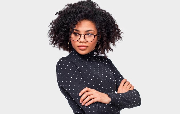 Photo portrait of serious african american young woman wears casual outfit and transparent eyeglasses afro female in black with white dots long sleeve shirt posing over white wall people and emotions