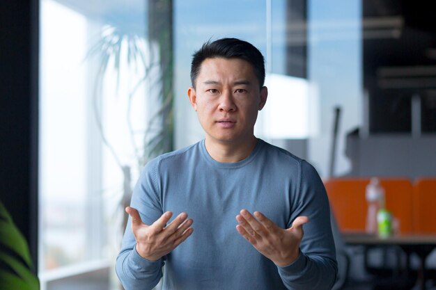 Portrait of a serious and angry employee looking at the camera and gesturing with his hands Asian communicates on a video call looks at the webcam