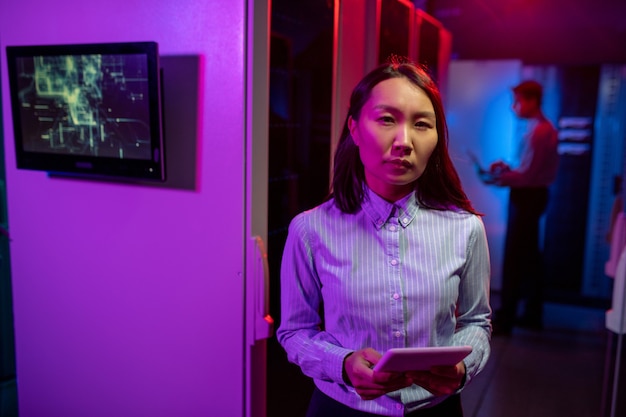 Портрет серьезной азиатской женщины-администратора сети, стоящей с планшетом против компьютерных шкафов в серверной