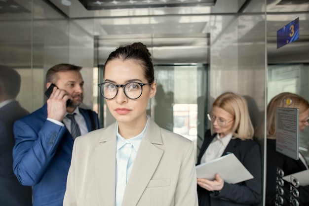 Portrait of serious attractive business lady standing in elevator with busy people in background
