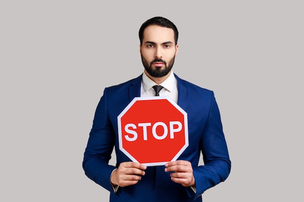 Portrait of serious bearded businessman holding Stop road traffic sign as symbol of prohibition, restrictions, wearing official style suit. Indoor studio shot isolated on gray background.