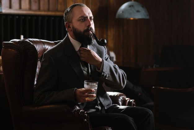 Portrait of serious bearded man with pipe holding glass of\
whiskey wearing suit and sitting on a big arm chair