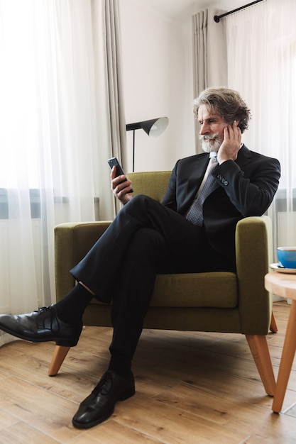 Portrait of a serious bearded mature grey-haired businessman posing indoors at home near window dressed in formal clothes using mobile phone sitting on chair.