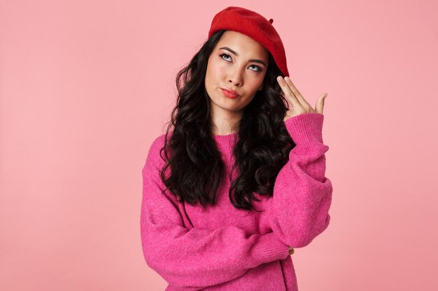 Portrait of serious beautiful asian girl wearing beret gesturing gun fingers at his temple isolated on pink