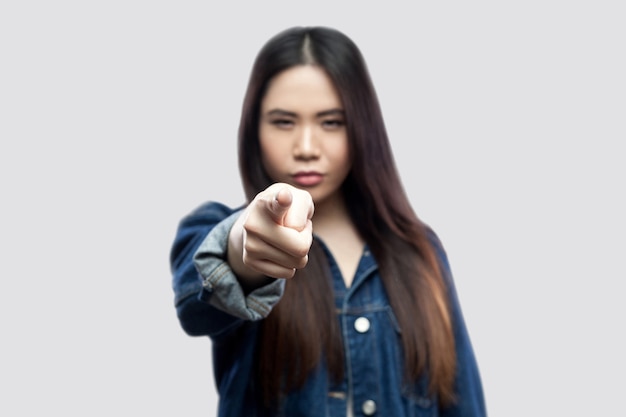 Portrait of serious beautiful brunette asian young woman in casual blue denim jacket with makeup standing pointing and looking at camera. indoor studio shot, isolated on light grey background