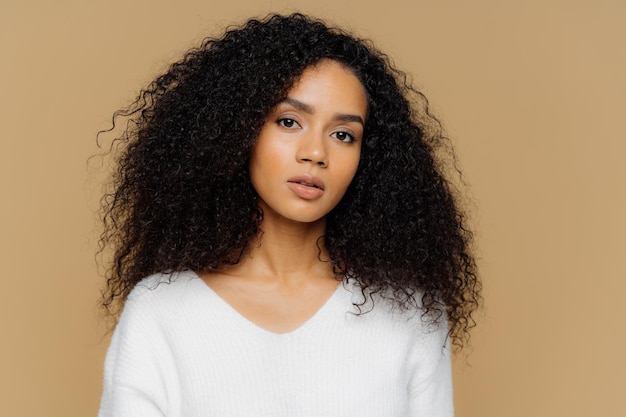 Portrait of serious beautiful dark skinned female with frizzy black hair has minimal makeup looks calmly at camera wears white jumper stands against brown background being deep in thoughts