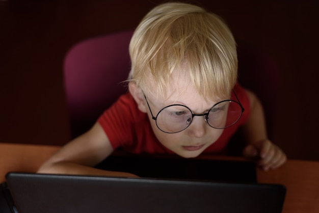 Portrait of a serious blond boy with glasses using a laptop