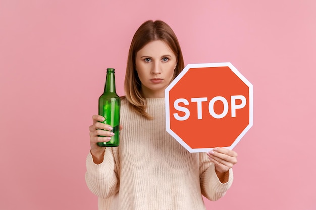 Portrait of serious blond woman holding red stop sign and bottle with alcoholic beverage calls on dont drink alcohol wearing white sweater Indoor studio shot isolated on pink background