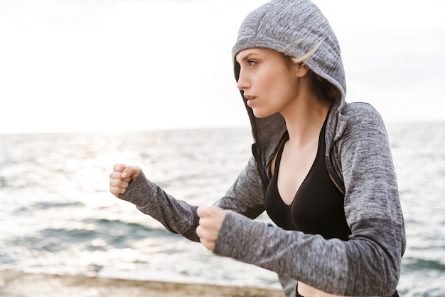 Ritratto di donna bionda seria che indossa la boxe con cappuccio mentre si allena sul molo vicino al mare al mattino