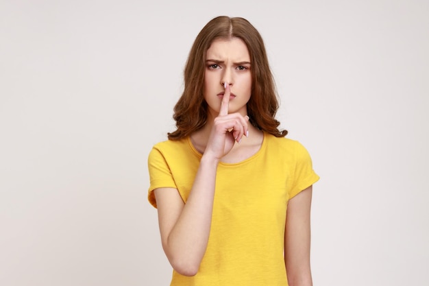 Portrait of serious brown haired teenager female in yellow casual T-shirt holding finger on lips, making hush silence gesture, asking to keep secret. Indoor studio shot isolated on gray background.