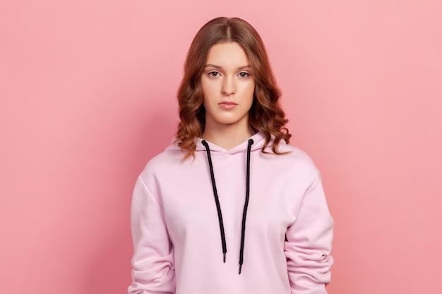 Portrait of serious brunette teenager girl in hoodie attentively looking at camera. Indoor studio shot, isolated on pink background