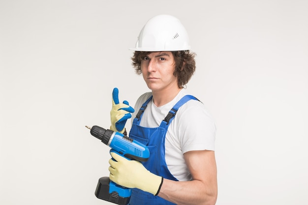 Портрет серьезного человека строителя в белом шлеме с перчатками отвертки и синими комбинезонами.