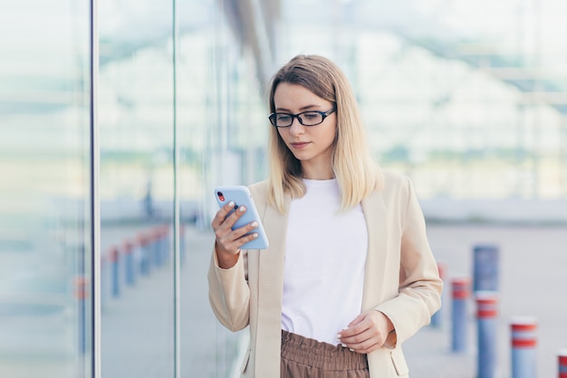 Портрет серьезной деловой женщины в очках, блондинка читает новости с мобильного телефона, использует приложение для перезаписи сообщений