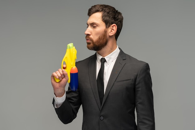 Портрет серьезного бизнесмена, играющего с водяным пистолетом и дующего в бочку со спокойным лицом на сером фоне