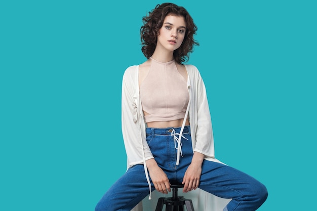 Portrait of serious calm beautiful young brunette woman in casual style sitting on chair and looking at camera with serious face. indoor studio shot, isolated on blue background.