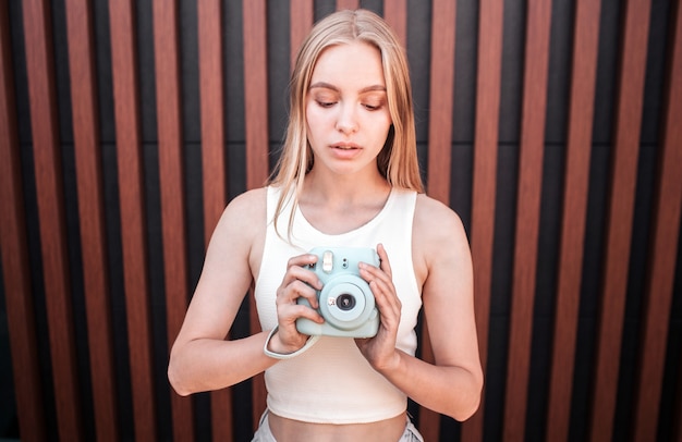 Portrait of serious and calm girl standing and holding blue camera. She is looking at it.