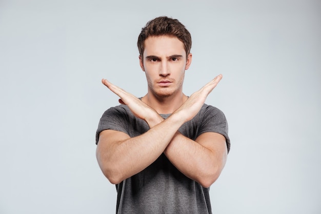 Portrait of a serious casual man showing crossed arms in a stop gesture on the gray