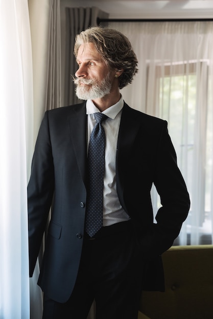 Portrait of a serious concentrated bearded mature handsome grey-haired businessman posing indoors at home near window dressed in formal clothes.