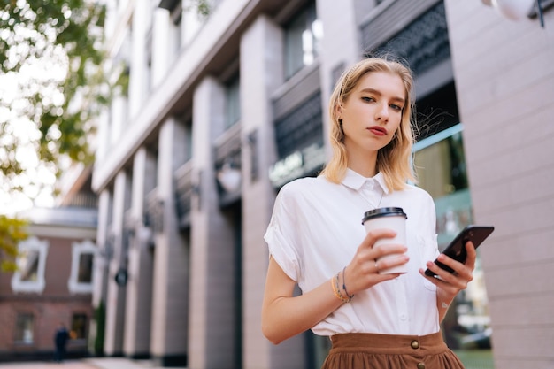 カジュアルな服を着て、持ち帰り用のコーヒーを飲み、カメラを見ながら携帯電話を使用する、真剣に自信を持っている金髪の女性ティーンエイジャーのポートレート