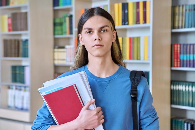 Ritratto di ragazzo serio studente fiducioso con libri di testo che guarda l'obbiettivo nella biblioteca del college