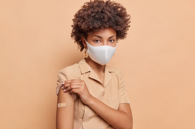 Portrait of serious curly haired woman rolls up sleeve of dress shows vaccination place wears adhesive bandage on arm recieves antivirus vaccine to safe life wears protective face mask poses indoor