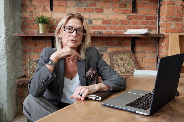 Portrait of serious fashionable mature business lady in jacket sitting at desk with open laptop in own office