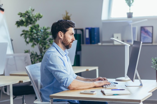 Foto ritratto di un uomo d'affari serio e concentrato, un uomo che lavora in un ufficio luminoso durante il giorno al computer guardando il monitor in abiti casual