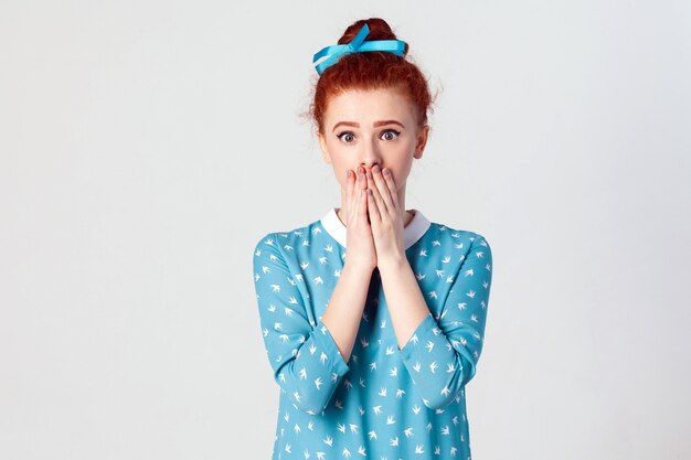 Portrait of serious girl covering mouth with both hands keeping a secret. Beautiful redhead female in blue dress doesn't want to spread rumors or some confidential information.